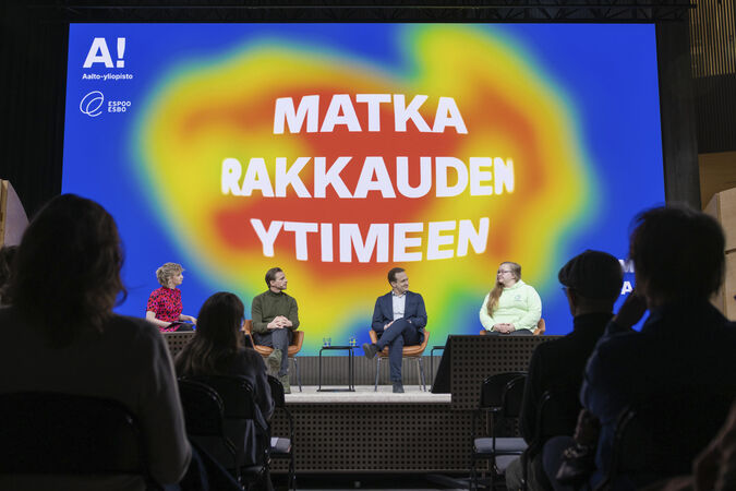 Neljä ihmistä istuu lavalla värikkään taustan edessä, jossa on suomenkielistä tekstiä. Yleisön jäsenet istuvat etualalla.