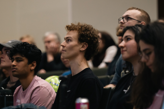 Students of varied looks sit closely in a lecture hall, watching a presentation with focused expressions.