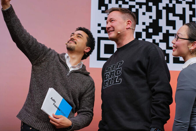 Three people on stage taking a selfie in front of a large black-and-white QR code.