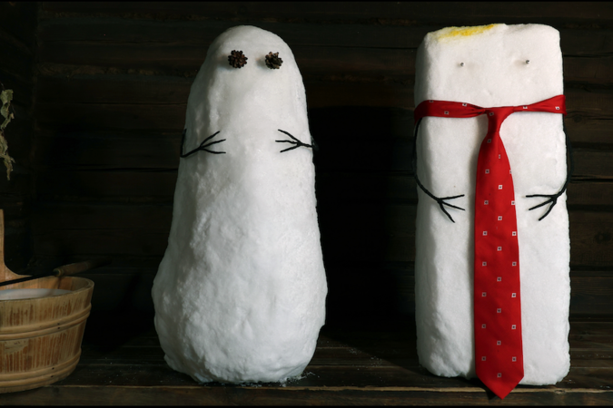 Two snow figures indoors, one plain with pinecone eyes, one tall with red tie, beside a wooden sauna bucket