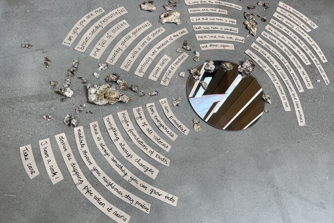 Brown paper text strips and stones spiral round a circular mirror on a smooth grey floor