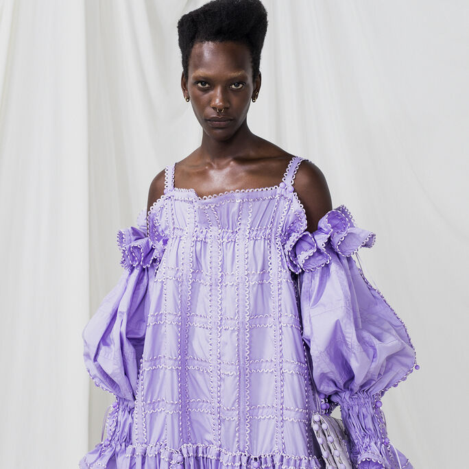 Person wearing a light purple dress with puffed sleeves and intricate patterns, holding a matching clutch.