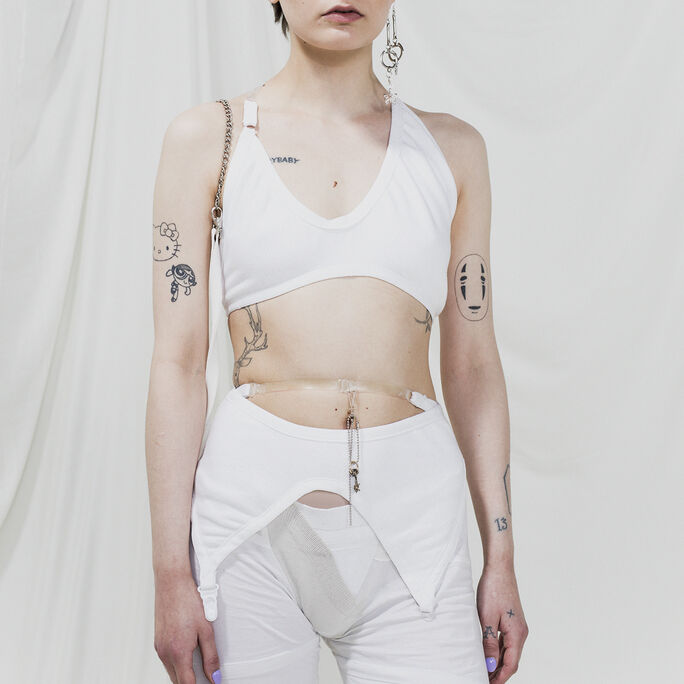 Person with short hair wearing a white crop top and pants, with multiple tattoos on arms and torso.