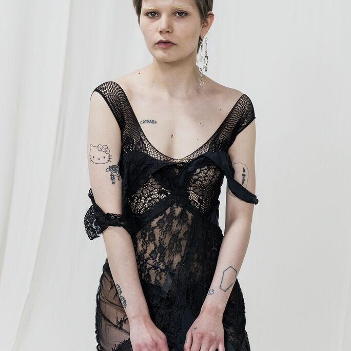 Person with short hair wearing a black lace dress, showing tattoos on arms and chest, sitting against a white background.