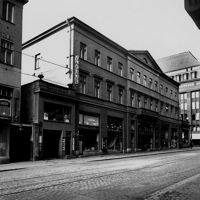 Mustavalkoinen kuva kadusta, jossa vanhoja rakennuksia, taustalla Stockmannin tavaratalo.