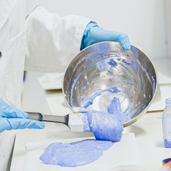Person in lab coat and blue gloves pouring blue paste from a metal bowl onto a white surface.