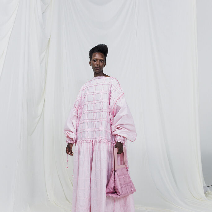 A person wearing a long, light pink dress with puffed sleeves, holding a matching pink handbag. White draped background.