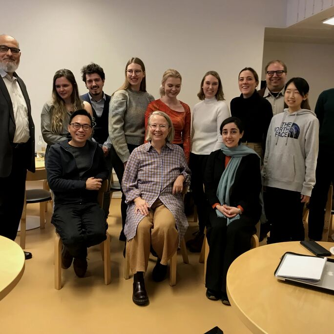 A group of people posing indoors, with one person seated in the centre. Tables and chairs are visible in the background.