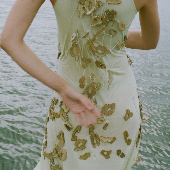 A person in a light green dress with floral patterns stands near a body of water, facing away.