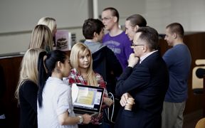 Two business students, one is holding a laptop, discussing with the Professor after his lecture. Another group of students talk in the background.