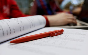 An Aalto pen lying on the page of a study book, students working in the background / photo by Aalto University, Aino Huovio