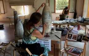 Kaksi naistaiteilijaa luomassa keramiikkataidetta Venetsian tutkimuspaviljongissa. Yksi istuu kuvan etualalla tuolilla ja tekee korkeaa maljakon muotoista esinettä, jossa näkyy hennosti naisen kasvot. Toinen henkilö on kuvassa taaempana, vähän kumartuneena, luoden isoa kulhoa.