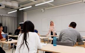 Opetus on käynnissä luokkahuoneessa ja opiskelijat kuuntelevat pöytään nojaavaa opettajaansa.