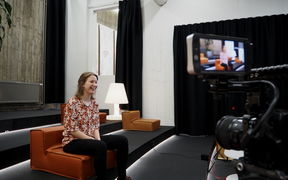 Riia-Leena Wallin sitting on a chair and laughing while being filmed on camera.