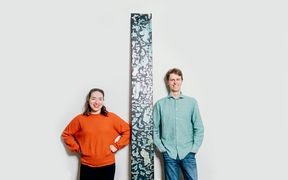 Tohtorikoulutettavat Noora Yau ja Konrad Klockars seisovat vierekkäin valkoista taustaa vasten. Heidän välissään on pystysuora puinen levy, joka on pohjaväriltään musta, mutta pinnassa on kirkkaita sinisiä ja keltaisia kuvioita. Kuva: Jaakko Kahilaniemi.