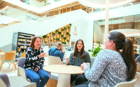 Three women are sitting at a cafe holding coffee cups and a bottle of water.