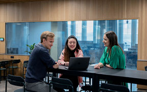 Two students and a professor sitting around a table, talking and looking at laptop screen.