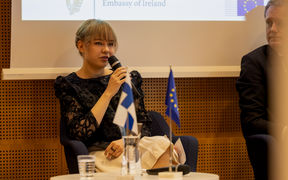 Ninni Norra puhuu nikrofoniin. Etualan pöydällä näkyvät pienet Suomen ja EU:n liput. 