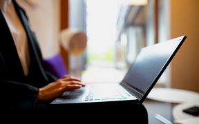 Henkilö kirjoittamassa kannettavalla tietokoneella, istuen ikkunan lähellä.