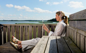 Henkilö istuu merinäköalan edessä kuulokkeet päässä ja selaa tablettia hymyillen.