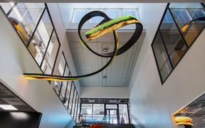 LED art portraying text in different colours, while meandering up in the ceiling and round a corner at K3 building. The picture is taken from ground floor towards the ceiling, showing the social stairs of the building and the second floor office glass walls.