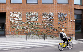 Punatiilisen rakennuksen julkisivussa on heijastavasta metallista tehty taideteos, jota katselee pyöräilijä.