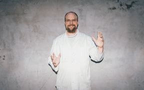 Valkoiseen laboratoriotakkiin pukeutunut Juho Uzkurt Kaljunen seisoo harmaan seinän edessä ja on levittänyt kätensä.