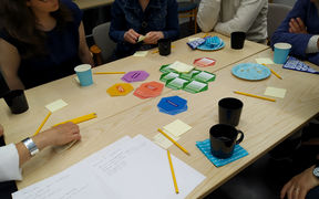 Ihmiset istuvat pöydän ääressä, jolla on papereita, kyniä ja värikkäitä kuusikulmiokortteja.
