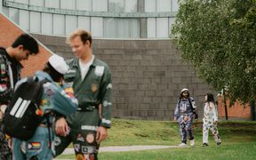 A group of people in colourful overalls with patches are gathered outdoors near a building and a tree.