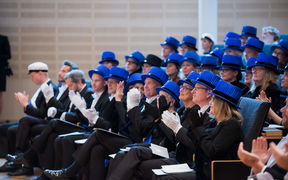 A group of people wearing formal attire and blue hats sit in rows, applauding at an indoor event.