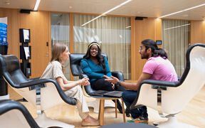 Three people sit in white chairs around a table with a laptop in a modern office space with wooden walls.