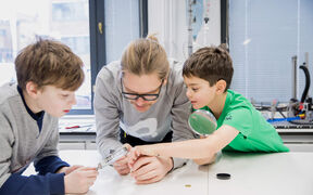 Kolme ihmistä tutkii esinettä pöydällä valoisassa huoneessa, jossa on suuret ikkunat ja tieteellisiä laitteita.