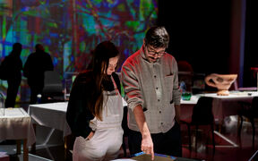 Kaksi henkilöä tarkastelemassa taidenäyttelyä. Mies osoittaa jotain pöydällä, huoneessa on erilaisia näyttelyitä värikkäällä taustalla.