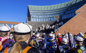 A student orchestra performs on steps while a crowd of students in colourful overalls and white caps watches.