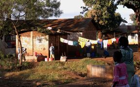 A rural scene with people washing clothes outside a small house. Colourful laundry hangs on a line. Trees and grass are visible.