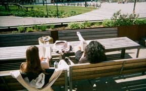 Two people are sitting on wooden benches outside, reading books. There is a grassy area and a bike park in the background.