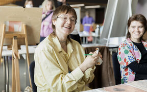 Henkilö keltaisessa paidassa pitelee valkoista figuuria ja vihreää kynää, istuu taiteen tarvikkeiden äärellä. Kuva: Aalto-yliopisto/Erica Nyholm