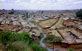 A view over densely packed houses in a slum area. Rusty metal roofs, scattered vegetation, and a narrow stream are visible.