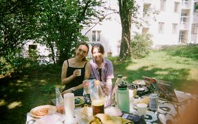 Kaksi henkilöä nauttii piknikistä puutarhassa, pöydällä on ruokaa, juomia ja kannettava tietokone.