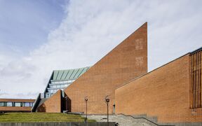 A modern building with sharp, angular designs in red brick and glass, under a cloudy sky.