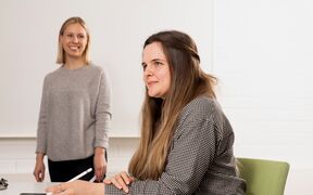 Kaksi hymyilevää henkilöä, joista toinen kuuntelee seisten ja toinen istuu kynä kädessä ja puhuu.