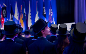 Graduates wearing black hats at a ceremony with flags in the background on a blue-lit stage.