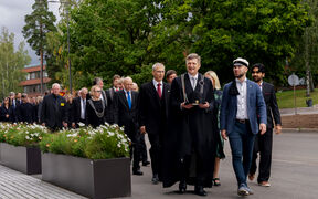 Aalto-yliopiston rehtori ja professoreiden kulkue muodollisissa vaatteissa kävelee ulkona ohi suurten istutuslaatikoiden, joissa on valkoisia kukkia. Taustalla puita ja rakennuksia.