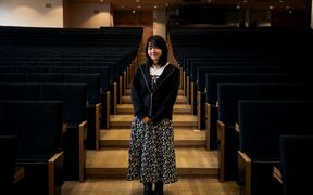 A person standing in an empty auditorium wearing a floral dress and black jacket with wooden seats.