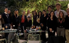 A group of people gathered outdoors at night. They are standing around tables with drinks, under trees and string lights.