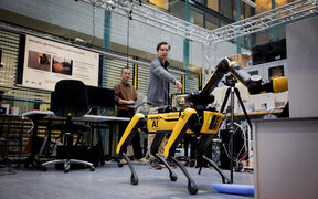 Two people interacting with a yellow Boston Dynamics robot dog in a tech lab with screens and equipment.
