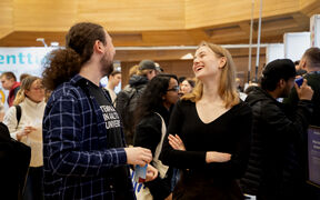 Kaksi ihmistä juttelee sisätapahtumassa. Yhdellä on ruudullinen paita ja kuppi, toisella musta paita ja kädet ristissä.