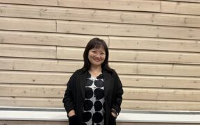 A person stands against a wooden wall, wearing a black jacket and a white shirt with large black dots.