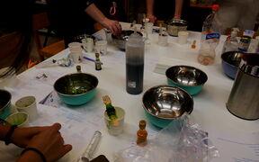 A table with bowls, bottles, and cups. People are working with various items, including a bottle of dark liquid.