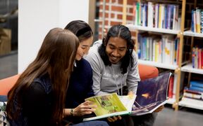 Three people sitting on a sofa, looking at a book together in a library with shelves of books in the background.